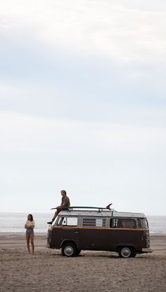 two people standing on top of an old vw van at the beach with surfboards