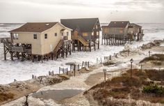 several houses on stilts in the ocean with waves crashing over them and sand covering the ground