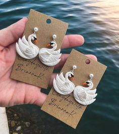 two white swan earrings are being held by someone's hand near the water with their name written on them