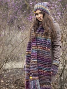 a woman wearing a multicolored scarf and hat standing in front of purple flowers