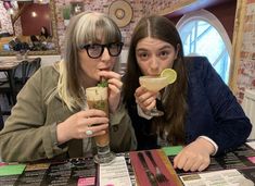 two women sitting at a table with drinks in front of their faces and menus on the table