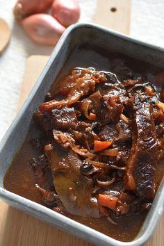 a pan filled with meat and vegetables sitting on top of a wooden table next to onions