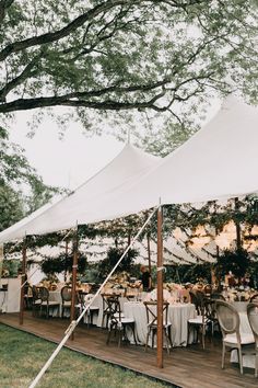 a large tent set up with tables and chairs for an outdoor wedding reception under the shade of a tree