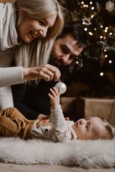 a man and woman playing with a baby in front of a christmas tree