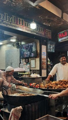 two men are cooking food at an outdoor stand in front of a restaurant with people standing around