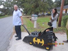 two people standing next to a batmobile on the side of the road, with one person holding a bat