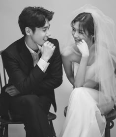 a man and woman sitting next to each other in front of a white wall wearing wedding veils