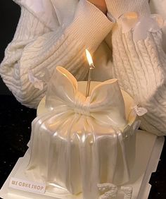 a candle that is in the middle of a cake with a bow on it and someone's hands behind it