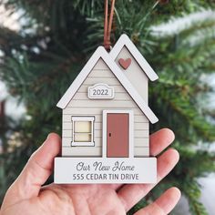 a hand holding a small house ornament in front of a christmas tree that says our new home 55 cedar tin drive