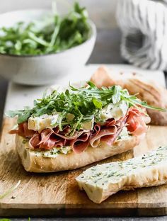 a wooden cutting board topped with a sandwich next to a bowl of greens and bread