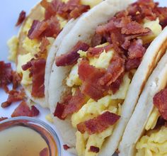 three breakfast tacos with eggs and bacon on them next to a cup of syrup