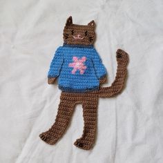 a crocheted cat wearing a blue sweater on top of a white bed sheet