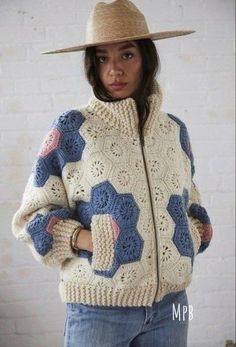 a woman wearing a hat and jacket with crocheted patterns on it, standing in front of a white brick wall