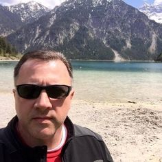 a man wearing sunglasses standing in front of a mountain lake