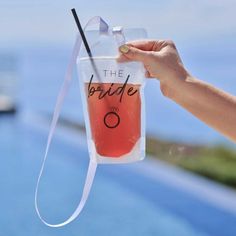 a hand holding up a plastic drink bag with the bride written on it next to a swimming pool