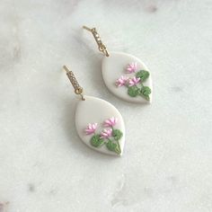 two white earrings with pink flowers and green leaves hanging from them on a marble surface