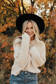 a woman wearing a black hat and sweater posing for a photo in front of trees