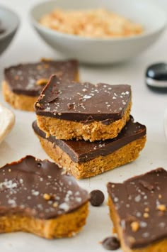 several pieces of chocolate and peanut butter bars on a white surface with bowls of cereal in the background