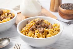 two white bowls filled with pasta and meat on top of a table next to silverware