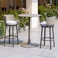 two stools and a table on the sidewalk near some plants in front of a building