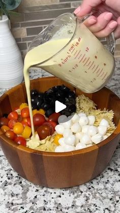 someone pouring dressing into a wooden bowl filled with fruit and veggie salad ingredients