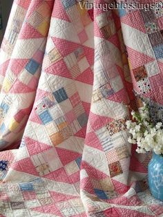 a pink and white quilt on a bed next to a blue vase with flowers in it