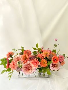 an arrangement of pink and orange flowers in a clear vase on a white cloth background