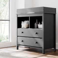 a gray dresser with two drawers in front of a white wall and window, next to a rug on the floor