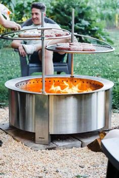 a man sitting in front of an outdoor fire pit with pizzas on the grill