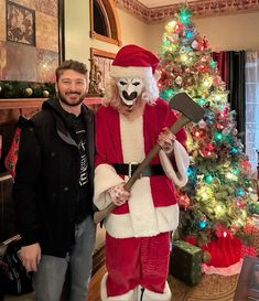 a man standing next to a person in a santa suit holding an ax near a christmas tree