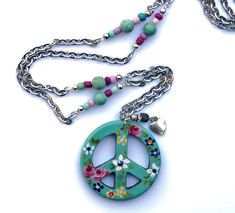 "A large turquoise blue peace sign pendant with colorful painted flowers in pink, yellow, and purple, dangles from a chain that's accented with genuine magnesite beads and little multi-colored wood beads! Sparkling Swarovski crystal rhinestones accent the pretty flowers and add a little glam! Dangling alongside the artful pendant, a little silver heart charm dangles below a genuine turquoise nugget and a rhinestone-encircled bead, adding funky movement! Just choose your sweater chain length to a Bohemian Blue Necklaces For Spring, Handmade Hippie Jewelry For Spring, Spring Bohemian Turquoise Jewelry, Spring Multicolor Hand-painted Jewelry, Flowers Romantic, Peace Sign Necklace, Hand Painted Flowers, Painted Flowers, Sweater Chain