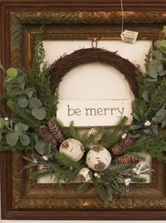 a wreath with pine cones and greenery is hung on the wall in front of a frame that says be merry