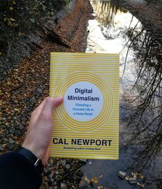 a person holding up a yellow book in front of a stream and trees with leaves on the ground