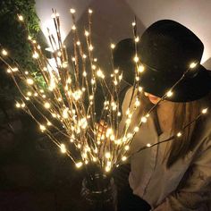 a woman wearing a black hat standing next to a vase filled with branches covered in lights