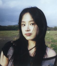 a woman with long black hair standing in front of an open field and looking at the camera