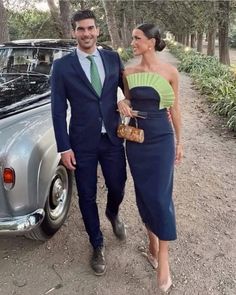 a man and woman standing next to a car in front of some trees on a dirt road