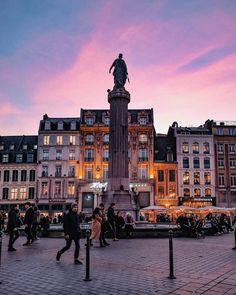 people are walking around in front of buildings at dusk, with a statue on top