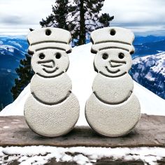 two snowmen made out of cement sitting on top of a snowy hill with mountains in the background