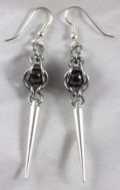 This product of Cassie's Creations is made from silver anodized aluminum rings that cage in a brown pearl bead, with silver spike charms that dangle down. The earrings are hung on black earring hooks. To get a list of the shows where we will be, visit www.MoonDragonDesigns.com, or message for custom orders Chain Maille Patterns Jewelry Making Tools, Caged Crystal Earrings, Chain Maille Patterns Earrings, Chain Mail Earrings Lone Rock Jewelry, Chainmail Patterns Earrings, Beaded Earrings Ideas, Chainmaille Jewelry Patterns, Jump Ring Jewelry, Black Earring