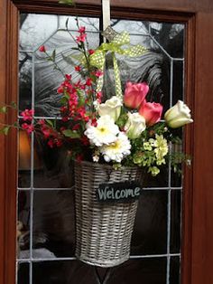 a welcome sign hanging from the side of a wooden door with flowers in a basket