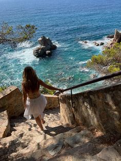 a woman walking up some steps towards the ocean with her hand on the railing and looking at the water