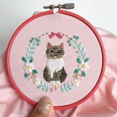 a cat is sitting in a floral wreath on a pink background with a hand holding the hoop