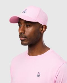 A person is wearing a pink MENS CLASSIC BASEBALL CAP - B6A816A1HT by Psycho Bunny and a matching pink T-shirt, both made of 100% cotton and featuring a small emblem of the Psycho Bunny logo with crossbones. The background is plain gray, and the person is looking slightly to the side with a neutral expression. Casual Visor Fitted Hat For Baseball Season, Casual Fitted Visor Hat For Baseball Season, Casual Fitted Hat For Sports Events, Casual Snapback Hat With Curved Visor For Baseball Season, Casual Trucker Hat For Sports Events In Spring, Casual Snapback Hat For Sports Events, Pink Hats For Spring Sports Events, Casual Summer Sports Fitted Hat, Casual Sports Baseball Cap With Curved Visor