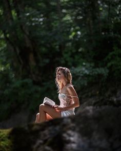 a woman sitting on a rock in the woods reading a book and looking at something