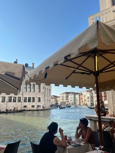 two women sitting at an outdoor dining area with umbrellas over looking the water and buildings in the background