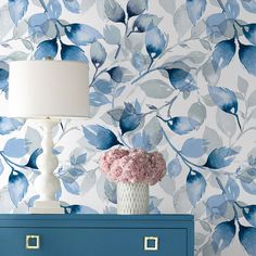 a blue dresser next to a white lamp and wallpaper with flowers on it,