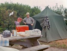 an old man standing in front of a table with boxes and bags on it next to a tent