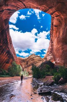 a man standing in the middle of a river under a large rock arch with a sky background