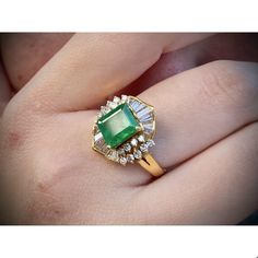 a close up of a person's hand holding a ring with an emerald and diamond