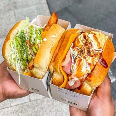 two hot dogs with toppings are in boxes on the street and someone is holding one